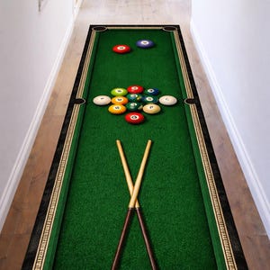 May include: A rectangular green rug designed to resemble a pool table, complete with billiard balls and crossed cues. The rug has a black border with gold accents and is placed on a wooden floor. The balls are arranged as if ready to be played.