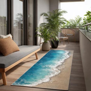 May include: A rectangular indoor/outdoor rug featuring a beach scene with blue ocean water and white foamy waves washing onto a sandy shore. The rug is placed on a wooden deck, with a gray chaise lounge and potted plants in the background.