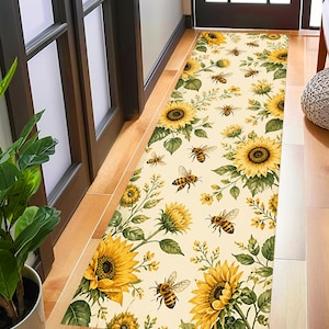 Può includere: Un tappeto rettangolare con un motivo ripetuto di girasoli gialli e api su sfondo crema. Il tappeto è posizionato in una porta, con pavimento in legno visibile su entrambi i lati. I girasoli sono in varie fasi di fioritura.
