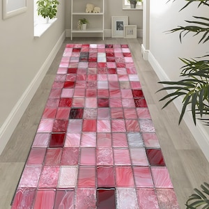 May include: A rectangular runner rug with a mosaic tile design in shades of pink and red. The rug features a repeating pattern of square tiles with varying textures and tones, creating a visually dynamic surface. The rug is placed on a light-colored wooden floor.
