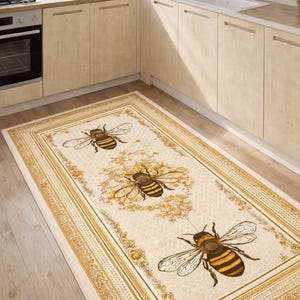 Op de afbeelding: Rechthoekig keukenkleed met een crèmekleurige achtergrond en een herhalend patroon van grote bijen. Het kleed heeft een gouden rand met bloemen en geometrische ontwerpen. De bijen zijn bruin en geel.