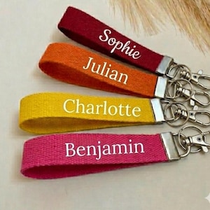 Peut inclure: Quatre porte-clés de poignet colorés : rouge avec "Sophie", orange avec "Julian", jaune avec "Charlotte" et rose avec "Benjamin". Chaque porte-clés a un fermoir argenté pour attacher aux clés ou aux sacs.