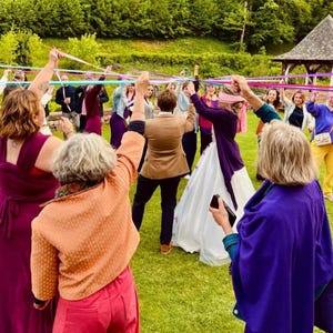 Peut inclure: Un groupe de personnes participe à une danse de la mai. Le mât de mai est orné de rubans colorés. Les participants portent diverses tenues, notamment des robes et des vestes. La scène se déroule sur une pelouse avec des arbres et un gazebo en arrière-plan.