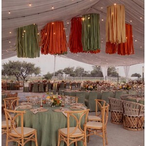 Pode incluir: Uma tenda para um evento ao ar livre, com mesas preparadas para uma celebra&ccedil;&atilde;o. As mesas s&atilde;o cobertas com toalhas verdes e decoradas com arranjos florais. Do teto pendem lanternas de papel nas cores verde, laranja e bege.