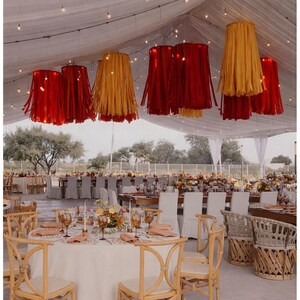 Pode incluir: Decora&ccedil;&atilde;o de recep&ccedil;&atilde;o de casamento com v&aacute;rias mesas e cadeiras sob uma tenda branca. L&acirc;mpadas com franjas vermelhas e amarelas penduradas no teto. As mesas s&atilde;o decoradas com centros de mesa florais, ta&ccedil;as e guardanapos.