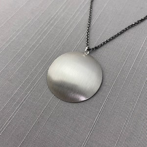 May include: A silver pendant necklace with a large, round, smooth, silver disc pendant. The pendant is attached to a thin, black chain.