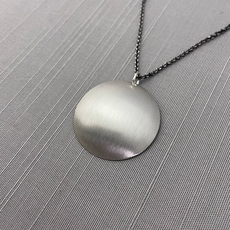 May include: A silver pendant necklace with a large, round, smooth, silver disc pendant. The pendant is attached to a thin, black chain.