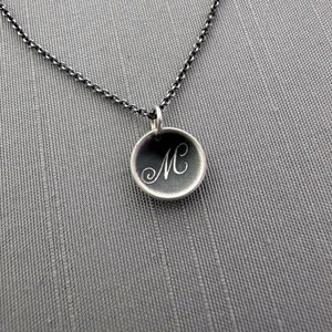 May include: A silver necklace with a round pendant featuring the letter "M" engraved in a cursive font.