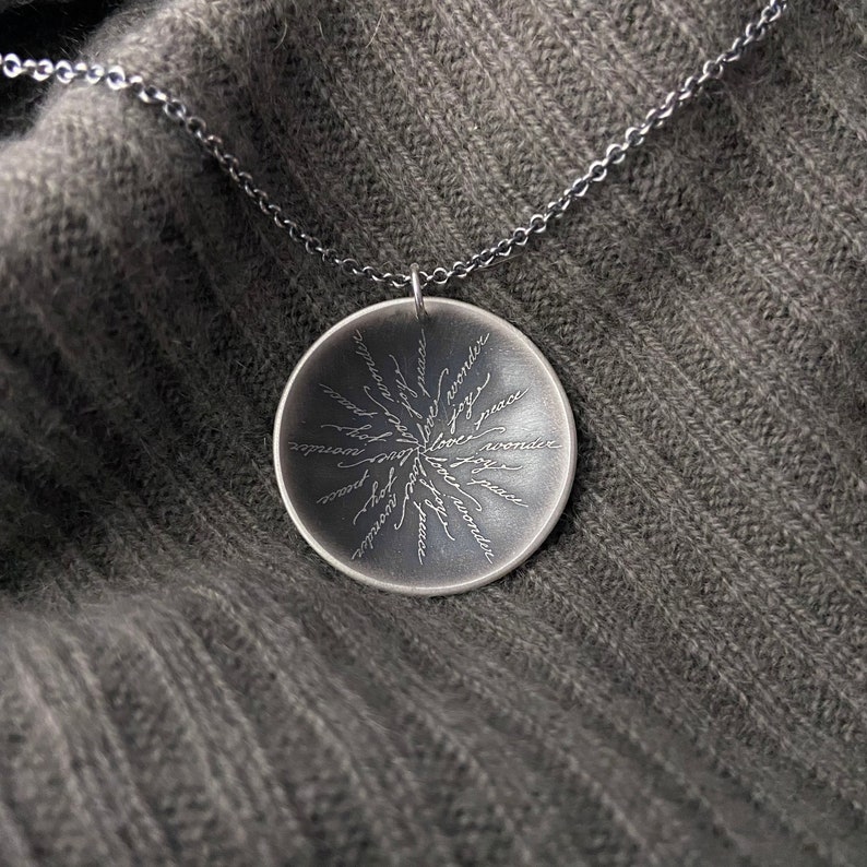 May include: A silver necklace with a round pendant featuring a circular design of words including "peace", "joy", "love", and "wonder".