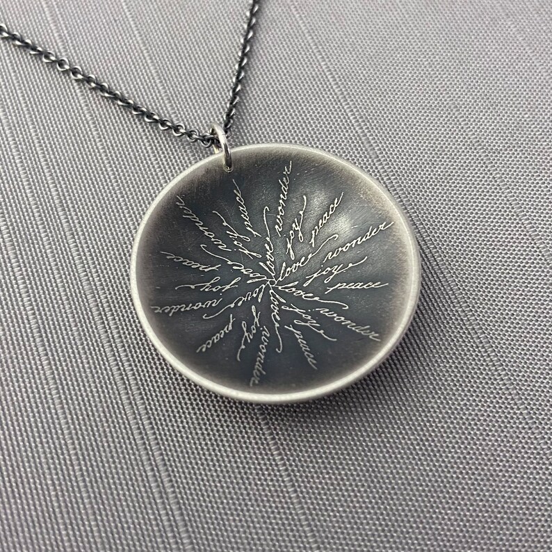 May include: A silver pendant necklace with a round disc featuring the words "love", "peace", "joy", "wonder", and "hope" inscribed in a circular pattern.