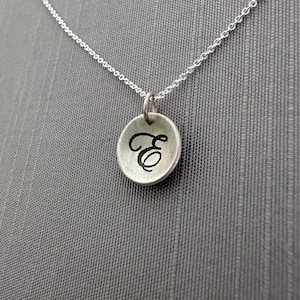 May include: A silver-toned necklace with a small, round pendant. The pendant features a black, cursive letter "E" in the center. The chain is delicate and the background is a solid gray.