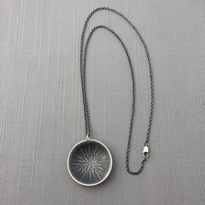 May include: A silver necklace with a round pendant featuring a detailed etched design. The pendant is about 2 inches in diameter and hangs from a silver chain.