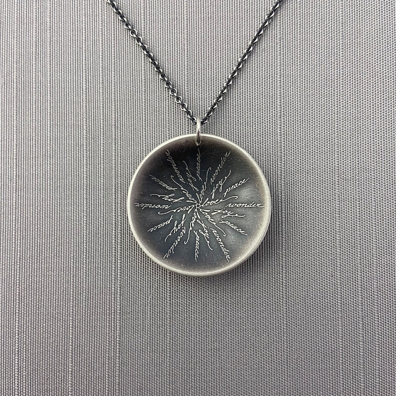 May include: A silver necklace with a round pendant featuring the words "joy", "peace", "love", "wonder", "hope", "faith", "gratitude", "compassion", "kindness", "strength", "wisdom", "courage", "inspiration", "joy", "peace", "love", "wonder", "hope", "faith", "gratitude", "compassion", "kindness", "strength", "wisdom", "courage", "inspiration" inscribed on it.
