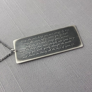 May include: A rectangular silver-toned metal pendant necklace featuring an engraved script message: May the sun bring you new energy by day, may the moon softly restore you by night, may the rain wash away your worries, may the breeze blow new strength into your being, may you walk gently through the world, and know its beauty all the days of your life.
