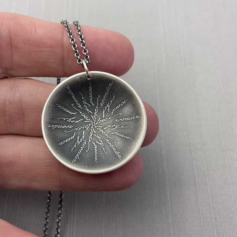 May include: Silver pendant necklace with a round disc featuring the words "love", "peace", "joy", and "wonder" inscribed in a circular pattern.