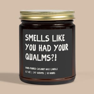 May include: A dark amber glass jar candle with a gold metal lid and a black label. The label features white text that reads: SMELLS LIKE YOU HAD YOUR QUALMS?! HAND-POURED COCONUT WAX CANDLE 8.7 OZ | 247 GRAMS | 50 HOURS. The candle is cylindrical in shape.