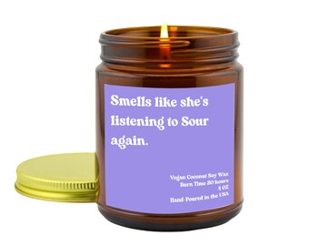 May include: A lit brown glass jar candle with a gold lid. The candle has a purple label with the text "Smells like she's listening to Sour again." The label also includes details about the wax, burn time, and size.