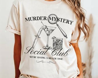 May include: A cream-coloured t-shirt with a black and white graphic. The design includes a martini glass with olives, a knife, a glass of whiskey, and the text "Murder Mystery Social Club" and "We're having a killer time."