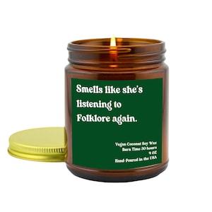 May include: A green glass candle jar with a gold lid. The label reads "Smells like she's listening to Folklore again."  Vegan Coconut Soy Wax, Burn Time 50 hours, 9 oz, Hand-Poured in the USA.