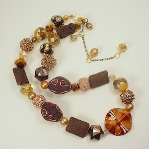 Puede incluir: Un collar de cuentas marrones, doradas y transparentes con una variedad de texturas y formas. El collar presenta una mezcla de cuentas redondas, cuadradas e irregulares, así como algunas con patrones intrincados. El collar está enhebrado en una cadena dorada.
