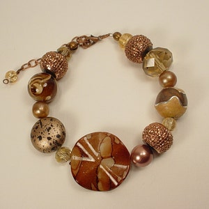 Puede incluir: Un brazalete de cuentas marrones y doradas con una cuenta grande, redonda, de color marrón y blanco con estampado en el centro.
