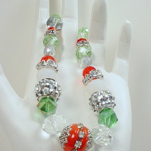 Puede incluir: Un brazalete de cuentas de colores con cuentas transparentes, verdes, rojas y plateadas. Las cuentas rojas tienen un diseño de pedrería. El brazalete se exhibe sobre una superficie blanca. Las cuentas se miden en centímetros.