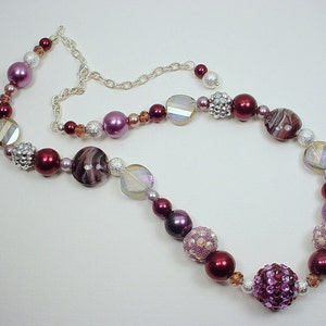 Puede incluir: Un collar largo de cuentas con una variedad de cuentas moradas, rosadas y plateadas. El collar tiene una cadena plateada y una gran cuenta morada brillante en el centro.