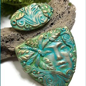 May include: Two decorative pieces featuring a teal and gold finish. The larger piece is a triangular plaque with a sculpted human face surrounded by leaves and a dragonfly. The smaller piece is an oval plaque with swirling leaf patterns.