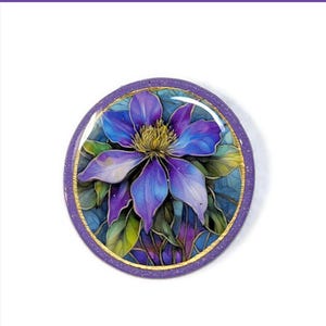 May include: A circular pin featuring a vibrant purple and blue clematis flower illustration. The artwork is encased in a glossy, clear dome with a glittery purple border and a thin gold-colored rim, showcasing detailed petals and green foliage.