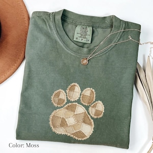 Puede incluir: Una camiseta Comfort Colors de color musgo con un diseño de huella de pata cosida. La huella de la pata presenta un patrón de patchwork en tonos beige y marrón. Un collar plateado con un colgante está drapeado sobre la camisa. El texto "Color: Moss" está en la parte inferior.