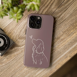 Puede incluir: Una funda de teléfono malva con un dibujo lineal blanco de un perro mirando un corazón. La funda tiene un acabado brillante y un recorte para la cámara. La funda del teléfono está sobre una superficie de madera.