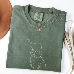 Puede incluir: Camiseta verde oliva Comfort Colors con un dibujo lineal blanco de un perro mirando un corazón. Un collar dorado con un colgante descansa sobre la camisa. La etiqueta dice "Comfort Colors".