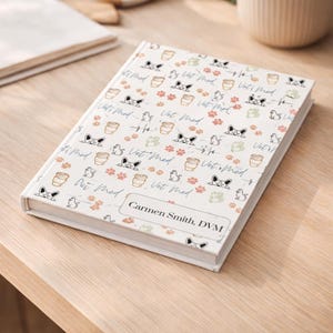 May include: A white notebook with a patterned cover featuring dog faces, paw prints, coffee cups, and the text "Vet Med." The notebook is personalized with the name "Carmen Smith, DVM."