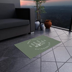 Könnte beinhalten: Rechteckiger, salbeigrüner Outdoor-Teppich mit dem Text "Happy Camper Outdoors Club" in Weiß. Das Design umfasst einen Camper, Bäume und einen kreisförmigen Rand. Der Teppich liegt auf einer gefliesten Oberfläche.