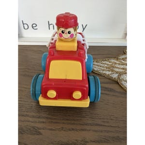 May include: A red and yellow toy fire truck with blue wheels and a smiling firefighter figure on top. The toy has the word "be happy" in the background.