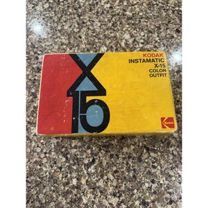 May include: A vintage Kodak Instamatic X-15 Color Outfit box. The box features a bold design with red, yellow, and blue colors. The Kodak logo is visible in the lower right corner. The box is rectangular and appears to be made of cardboard.