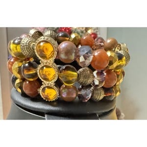 May include: A stack of beaded bracelets featuring a variety of amber, brown, and gold beads. One bracelet has three large amber-colored stones set in a gold-colored metal. Other beads are faceted, round, and textured.