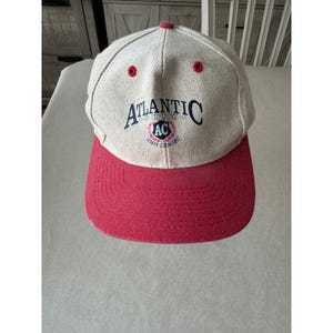 May include: A baseball cap with a red brim and a beige crown. The cap features the text "ATLANTIC" in navy blue, with "AC" in a circular emblem. Two red eyelets are on top.