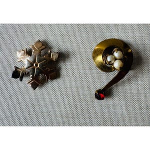 May include: Two vintage brooches. One is a gold-toned snowflake with six points. The other is a gold-toned swirl with three small pearls and a red gemstone at the end of the curved tail. Both are on a gray textured surface.