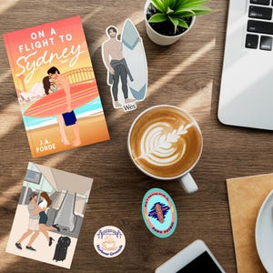 May include: A flat lay of a wooden table with a book titled "On a Flight to Sydney" by J.A. Forde, a coffee cup, a laptop, a sticker of a man on a surfboard, a sticker of a bridge with a coffee cup, and a sticker of a surfboard with the text "Dawn Patrol".