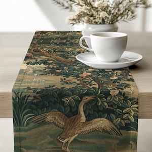 May include: A rectangular table runner featuring a dark green botanical print with a central brown goose with outstretched wings, surrounded by lush foliage, blooming flowers, and a fountain, with a white ceramic cup and saucer placed on top.