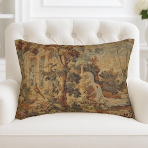 Puede incluir: Cojín decorativo rectangular con un diseño de tapiz que presenta una escena de paisaje con una cascada, árboles y un pájaro. El cojín tiene un fondo color crema y está colocado sobre un sillón blanco.