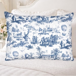 May include: A rectangular pillow with a blue and white toile pattern. The design features scenes of people, horses, trees, buildings, and hot air balloons. The pillow is placed on a white bed.
