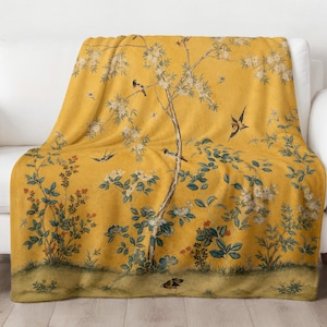 Puede incluir: Una manta de color amarillo mostaza con un estampado botánico detallado que muestra árboles esbeltos, flores blancas en flor y varios pájaros volando y posados en las ramas. El diseño incluye follaje verde y un pequeño pájaro en el suelo.