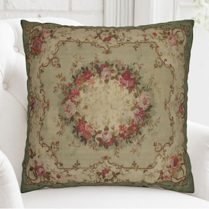 May include: Square cushion with a floral pattern. The cushion displays a central wreath of pink and red roses, set against a light green backdrop and a dark green border. The design has a vintage, antique aesthetic.