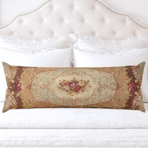 May include: A long, rectangular decorative pillow with a floral design. The pillow features a central oval with a bouquet of pink and red roses, surrounded by a beige and light green background. The edges are a darker brown with floral accents. The pillow is placed on a white bed.
