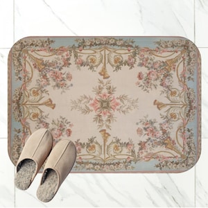 Puede incluir: Alfombra rectangular con un diseño floral en tonos crema, rosa y dorado, enmarcada por un borde azul claro. Dos zapatillas beige están colocadas sobre la alfombra. El patrón incluye rosas y elementos decorativos.