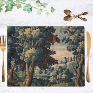 May include: Rectangular placemat featuring a landscape scene with trees and a body of water. The placemat is surrounded by a gold fork, knife, and spoons. Green ivy and a white wooden surface complete the scene.
