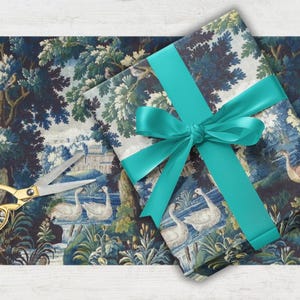 May include: Gift wrapping paper with a blue and white landscape design featuring swans, trees, and a building. A teal ribbon is tied around the wrapped gift. Gold scissors are next to the wrapping paper.