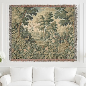 May include: A woven tapestry featuring a pastoral landscape with trees, flowers, and a deer. The tapestry has a fringed edge and is displayed on a white wall above a white sofa. The color palette includes greens, beige, and touches of pink.